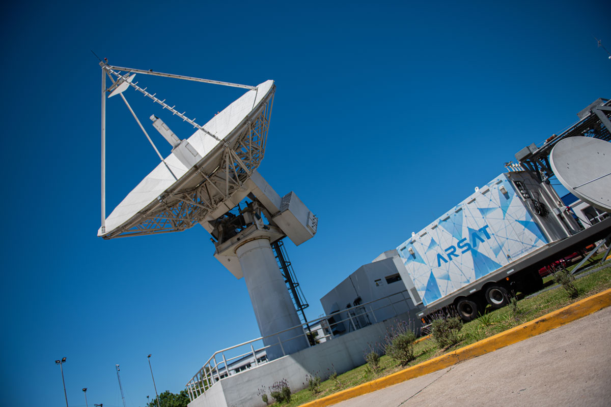 Una gran antena de la Estación Terrena de Benavídez, que apunta hacia el cielo donde están el Arsat 1 y 2, junto a un shelter con el logo de Arsat.